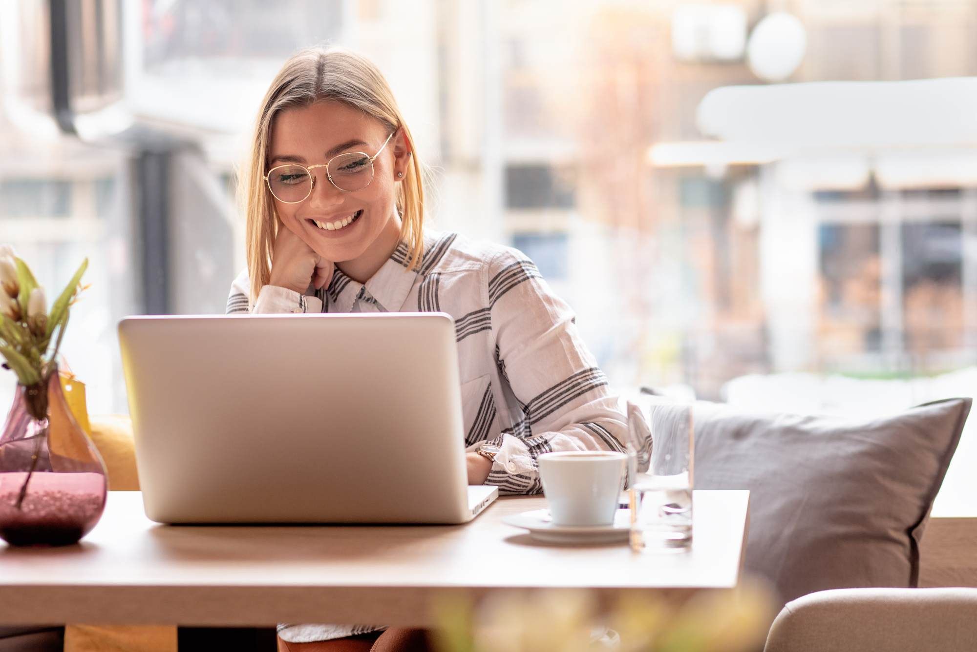 Smiling woman with glasses works on a laptop at a café table with coffee and a vase nearby. at Bluff View at Frederick Creek.