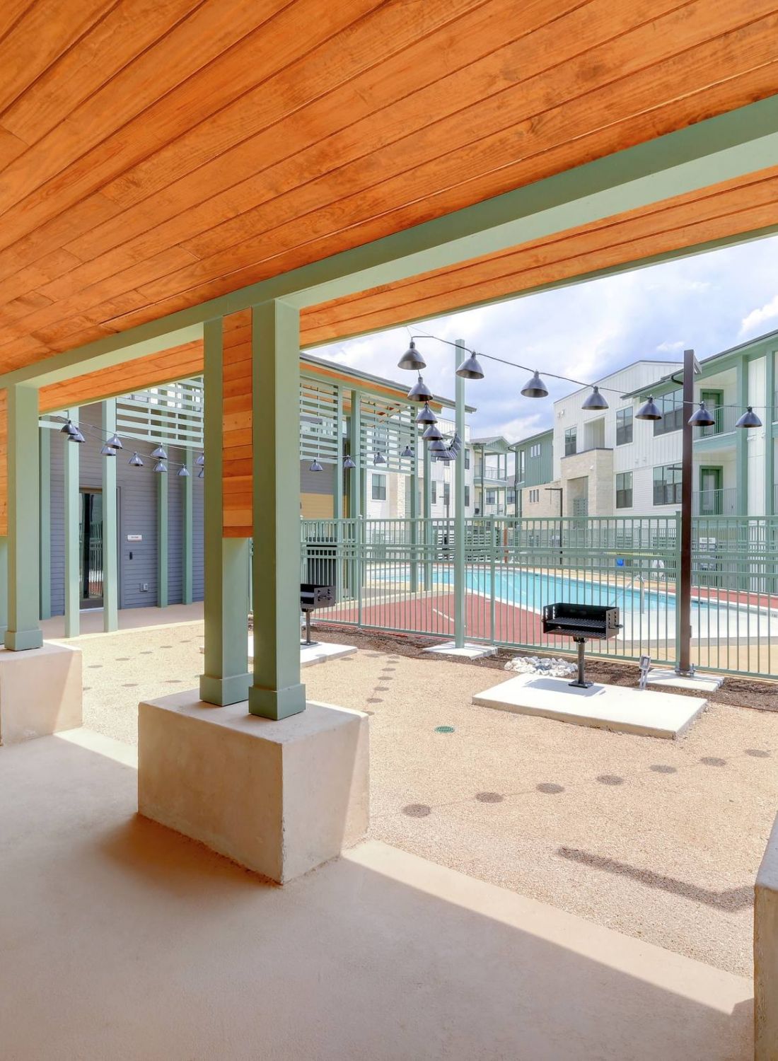 Covered patio area with green pillars overlooking a fenced pool and modern apartment buildings at Bluff View at Frederick Creek.