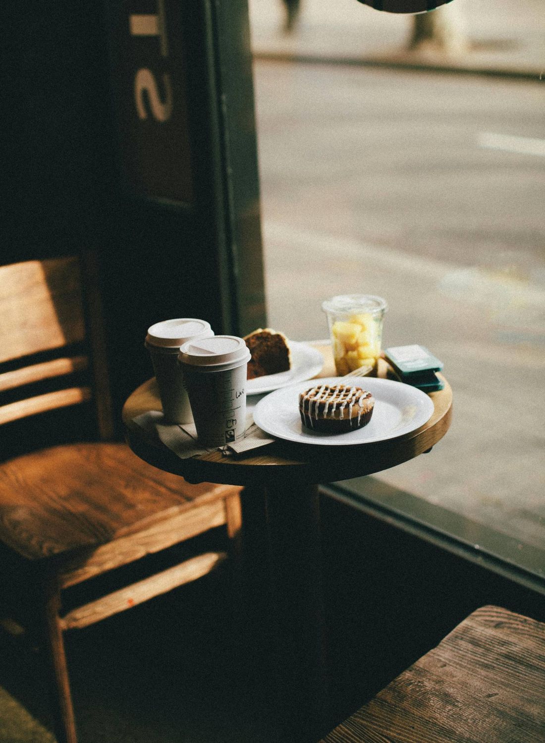 A small café table with two coffees, a pastry, fruit cup, and cake by a window with wooden chairs.
