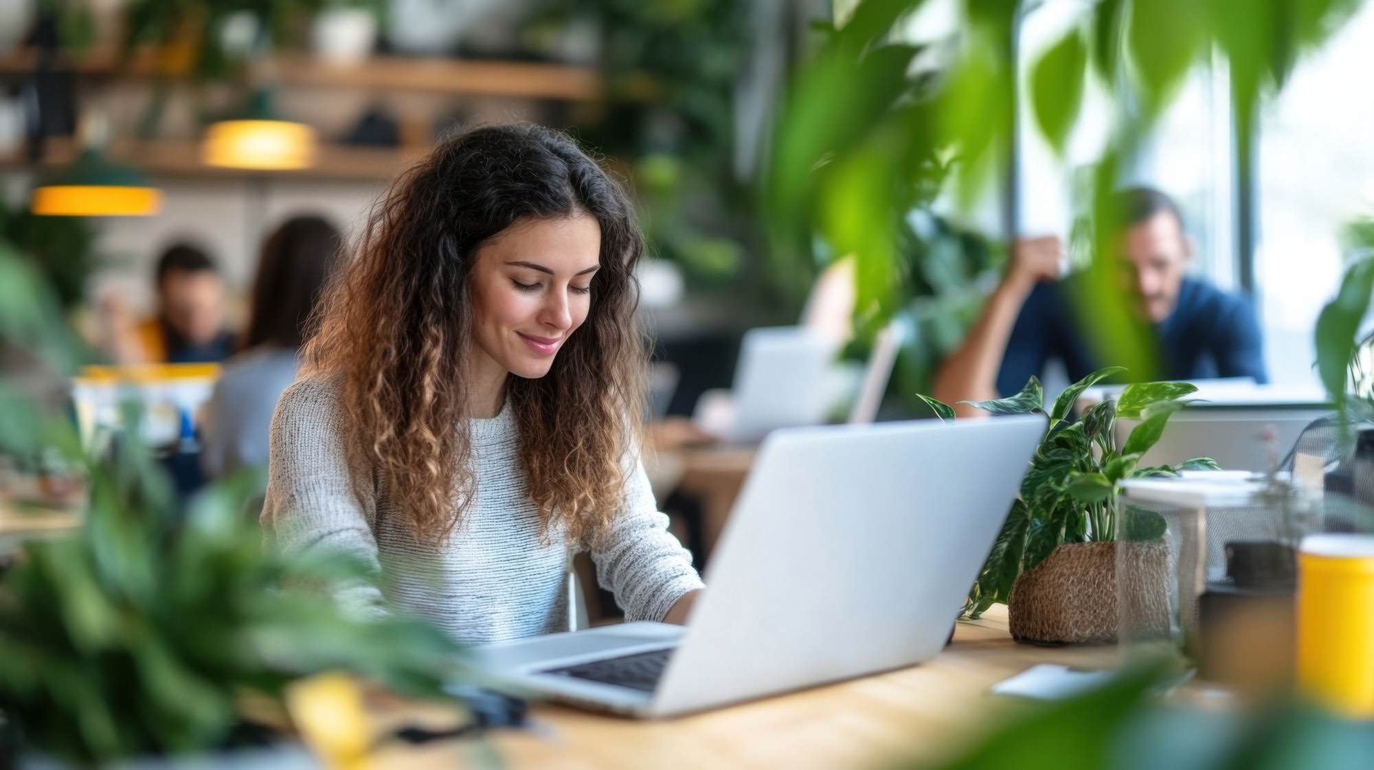 Woman with curly hair working on a laptop in a bright, plant-filled office space. at Bluff View at Frederick Creek.