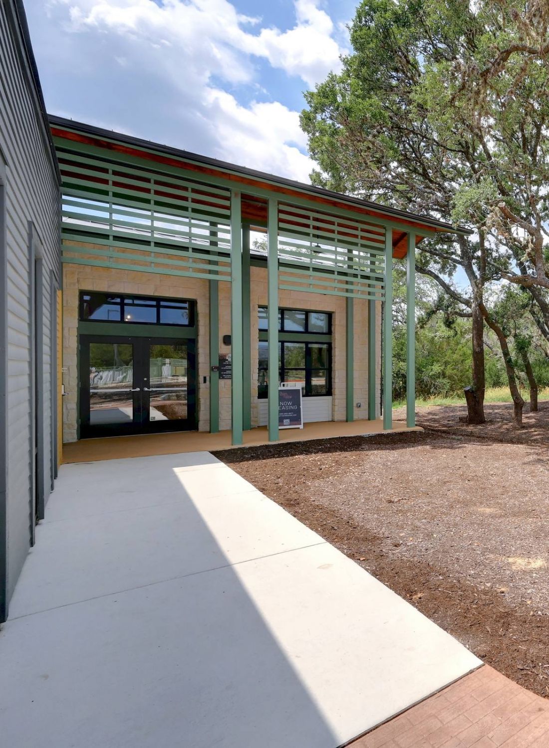 Modern building entrance beside large trees and a dirt area under a partly cloudy sky at Bluff View at Frederick Creek.