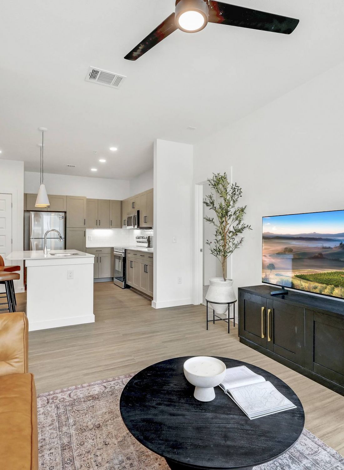 Modern living room with a TV, brown sofa, round coffee table, and open kitchen in the background at Bluff View at Frederick Creek.