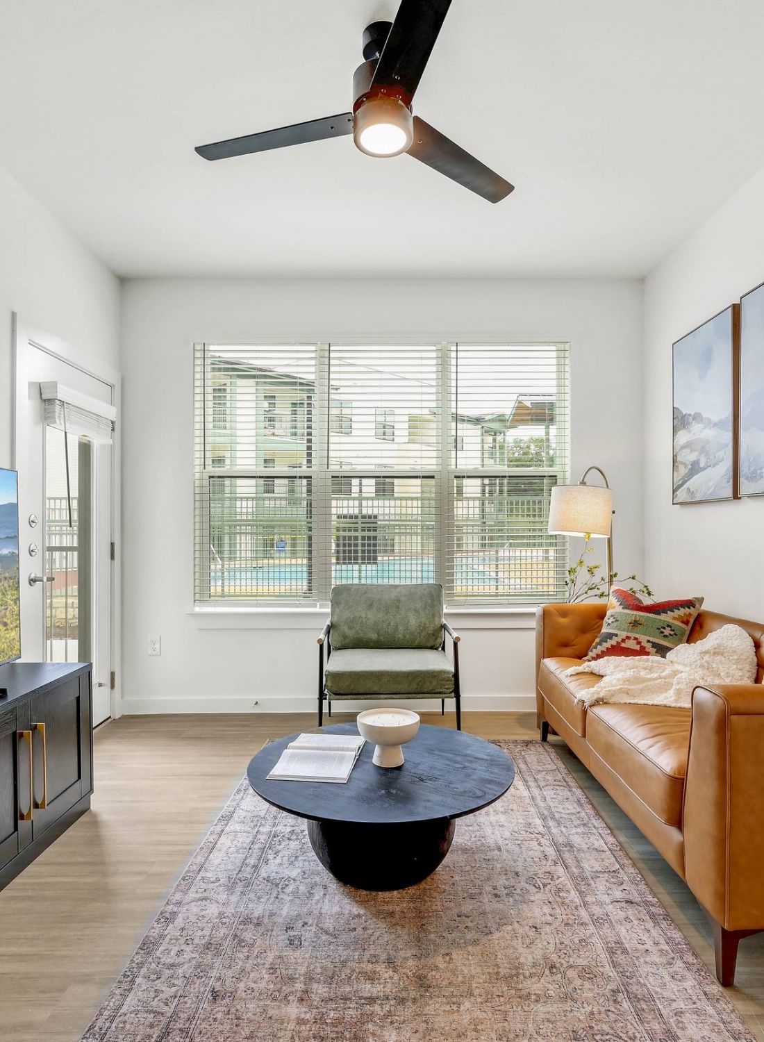 Modern living room with a tan leather sofa, TV, large window, art on the wall, and a round coffee table at Bluff View at Frederick Creek.