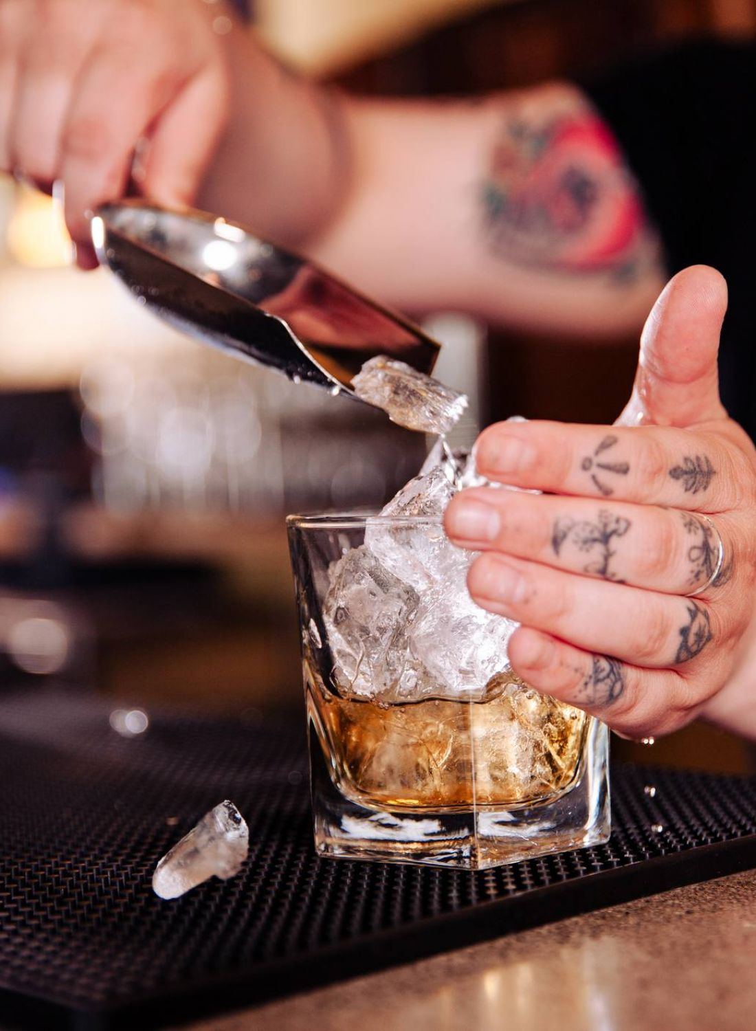 A bartender with tattoos adds ice to a glass of whiskey at a bar counter. 