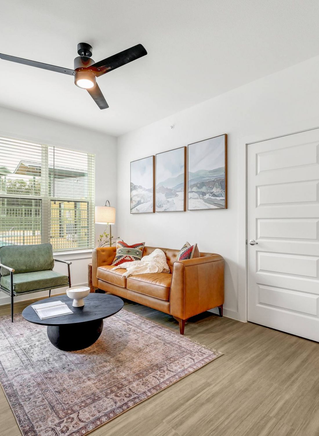 Modern living room with tan sofa, green chair, round coffee table, large window, and ceiling fan at Bluff View at Frederick Creek.